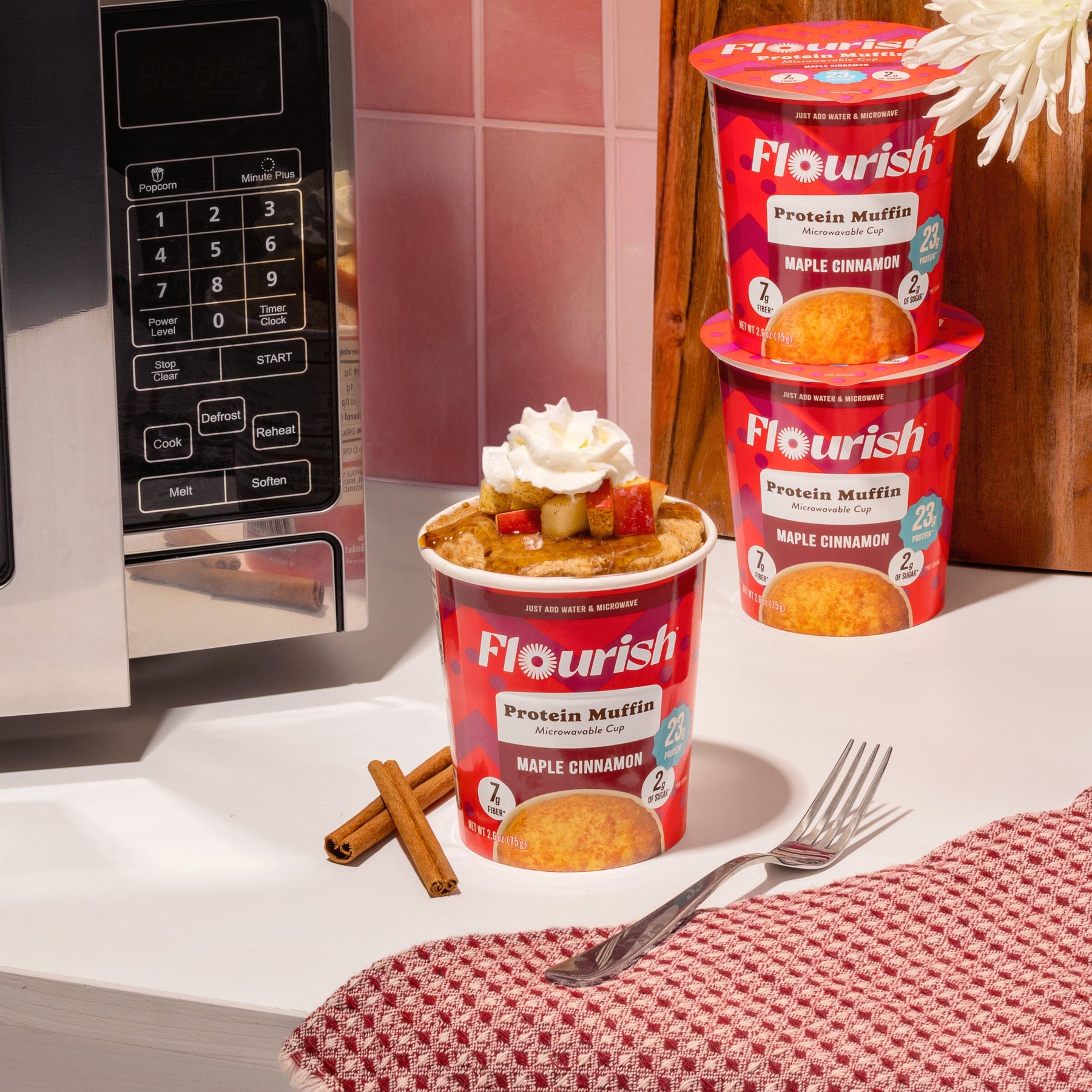 Prepared Flourish Maple Cinnamon Protein Muffin Cup topped with whipped cream, apples, and maple syrup beside a microwave, ready in seconds.